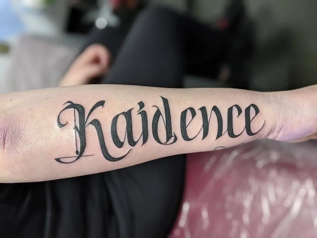 a black ink tattoo on a forearm with the name kaidence written in elegant cursive script against a light skin background with a soft bruise above it
